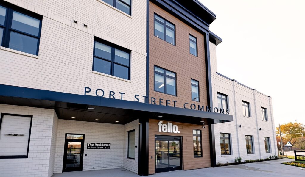 Exterior of a modern building named "Port Street Commons" with a sign for "Fello" near the entrance. The structure is three stories tall, featuring a combination of white brick and wood-like paneling, with multiple windows. There's a sign for "The Residence" above one of the doorways. The surrounding area is paved with some greenery visible and a clear sky above.